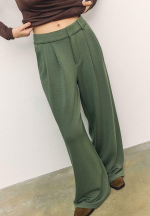 Person trägt hochgeschnittene, weit geschnittene olivgrüne Hose und braune Schuhe, hebt braunes Oberteil an, um den Bauch vor einem einfarbigen Hintergrund zu zeigen.