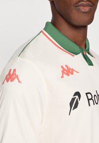 Kremo spalvos polo marškinėliai su žaliu apykakle ir raudonomis detalėmis. Ant rankovių yra rausvos „Kappa“ logotipai, o krūtinėje – juodas „Robor“ ženklinimas. Audinys su tekstūra.