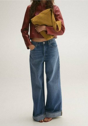 Personne portant un jean bleu à jambes larges avec des revers, une veste en cuir rouge courte, tenant un grand sac jaune moutarde, debout sur un fond uni.