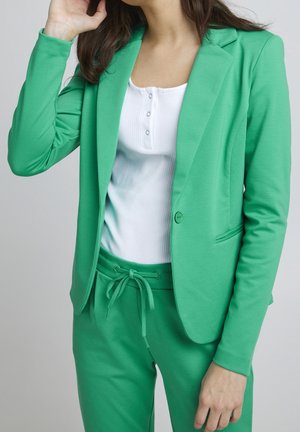 Personne portant un blazer vert vif et un pantalon assorti avec cordon de serrage, associé à un haut blanc côtelé à boutons, main près du visage.