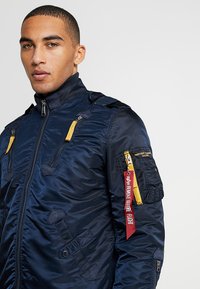 Navy blue bomber jacket with a high collar, multiple zippered pockets, yellow accents, and a red "Remove Before Flight" tag. Smooth texture.