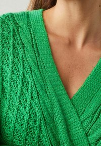 Nahaufnahme einer Person, die einen grünen Strickpullover mit Kabelmustern und V-Ausschnitt trägt.