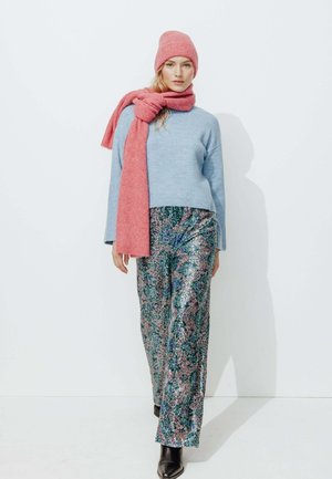 Pull bleu clair, bonnet et écharpe en tricot rose, pantalon large vert pailleté, bottines noires. Textures lisses avec des motifs vibrants.