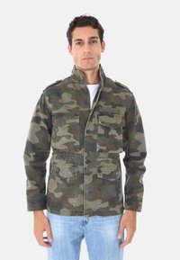 Giacca camouflage in tonalità verde e marrone, con colletto alto, numerose tasche e chiusura a bottoni. Indossata sopra una camicia bianca, abbinata a jeans.