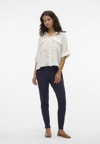 Biely košeľový tričko s dvoma prednými vreckami, vyhrnutými rukávmi, v kombinácii s tmavomodrými slim-fit nohavicami a hnedými balerínkami. Pozadie je jednoduché.