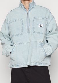 Ljust blå denimjacka med hög krage, halvdragkedja fram, två stora framfickor och en liten logopatch på vänster bröst.