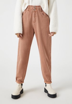 Pantalons marron taille haute avec poches avant, deux boutons, jambes droites et ourlets retroussés, associés à des bottines blanches à la cheville.