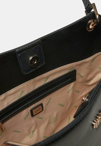 Interno di una borsa nera in materiale sintetico con una texture liscia, dotata di una tasca con zip e foderata in beige con loghi "GUESS" in verde.