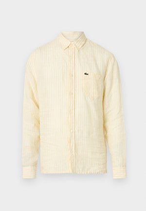 Camisa de manga larga con rayas amarillas y blancas, diseño con botones, con un bolsillo y un detalle de logo verde en el frente. Textura de tela suave.