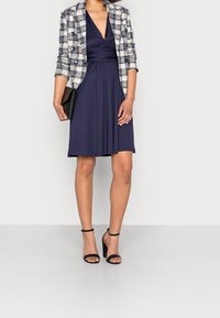 Marinblå klänning med v-ringning och rynkad midja, kombinerad med en rutmönstrad kavaj. Svarta remsandaler med klack och en liten svart clutch.