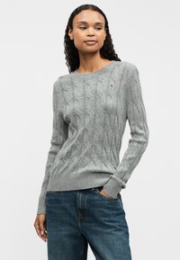 Grauer Grobstrickpullover mit geripptem Saum und Bündchen. Verfügt über ein kleines Logo auf der Brust. Kombiniert mit blauen Jeans, die eine lockere Passform zeigen.
