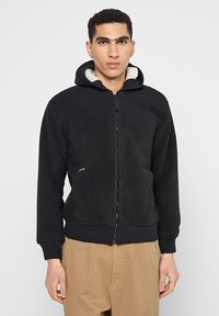Schwarze Kapuzenjacke mit Reißverschluss, innen weich und strukturiert gefüttert, mit gerippten Bündchen und Saum sowie einem kleinen Logodetail.