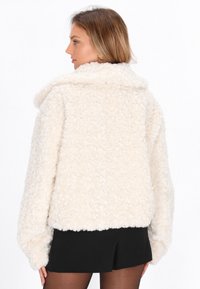 Vrouw in een crèmekleurige fluffy jas en zwarte shorts, zijwaarts gekeerd met ontspannen handen langs haar zij tegen een witte achtergrond.