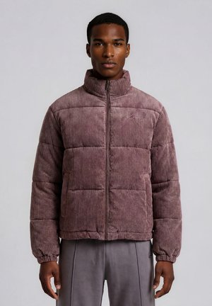 Jeune homme portant une veste matelassée en velours côtelé mauve avec un col montant et un pantalon gris, debout devant un fond uni gris clair.