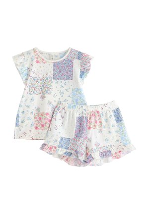 Lille barn med krøllet hår iført hvid blomstret patchwork top med flæseærmer og matchende shorts står barfodet på ensfarvet baggrund.