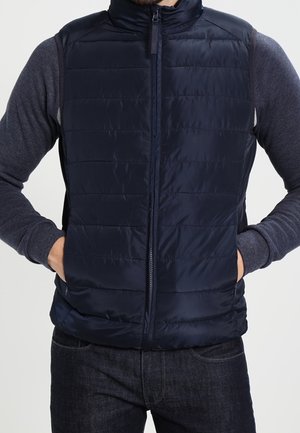 Homme portant un gilet matelassé bleu marine sur une chemise grise à manches longues, les mains dans les poches du gilet, montré des épaules jusqu’aux hanches.