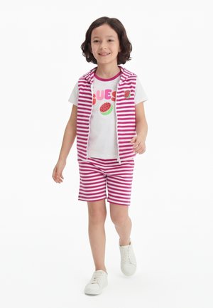 Μικρό παιδί περπατάει φορώντας λευκά αθλητικά παπούτσια, ροζ και λευκά ριγέ σορτς και γιλέκο με φερμουάρ, και λευκό μπλουζάκι με γραφικό φρούτου και κείμενο.