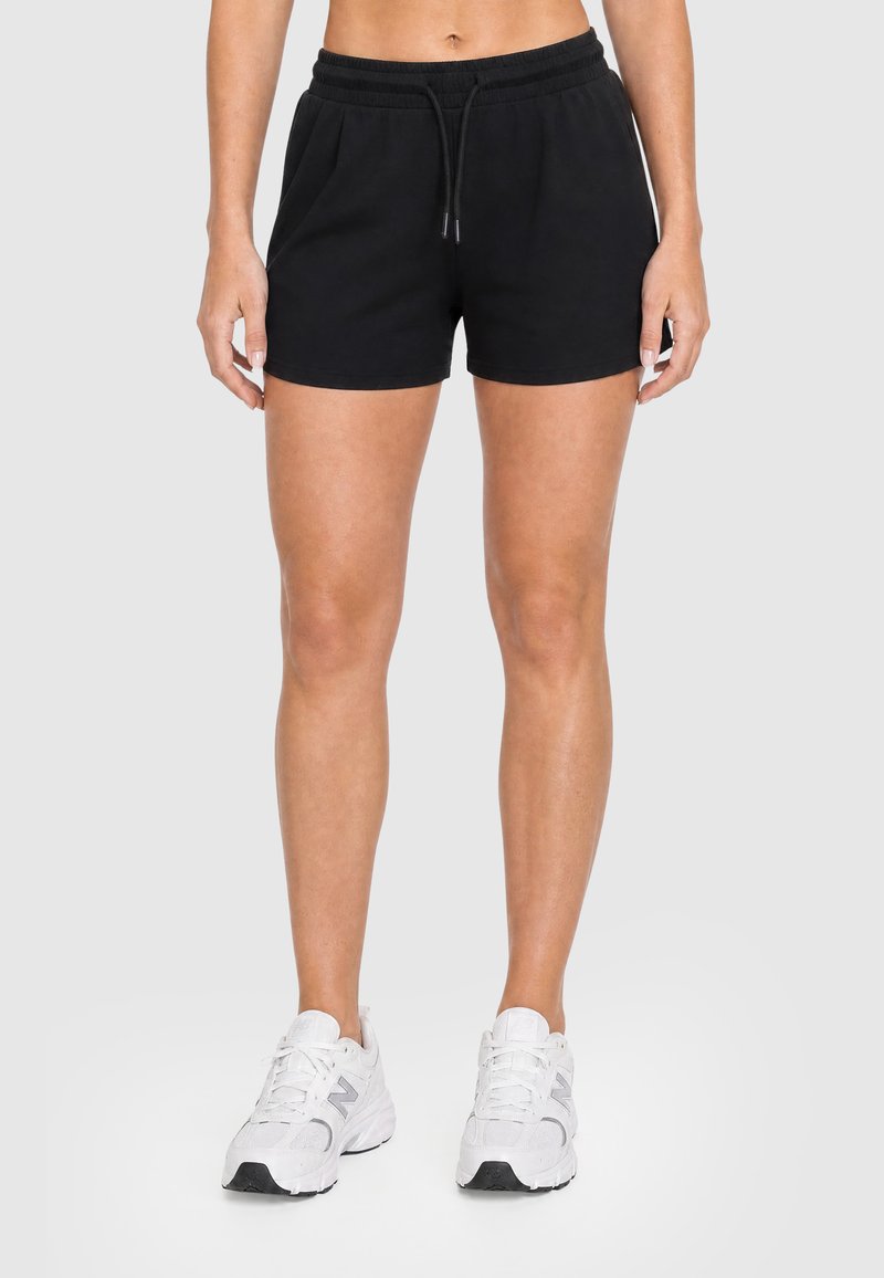Femme portant un short noir à taille élastique et des chaussures de sport blanches, debout devant un fond uni.