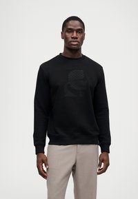 Μαύρη μπλούζα με κουκούλα που διαθέτει υφή στο γραφικό τύπωμα μπροστά, στρογγυλή λαιμόκοψη και ριμπ μανίκια. Το μοντέλο φοράει ανοιχτό γκρίζο παντελόνι.