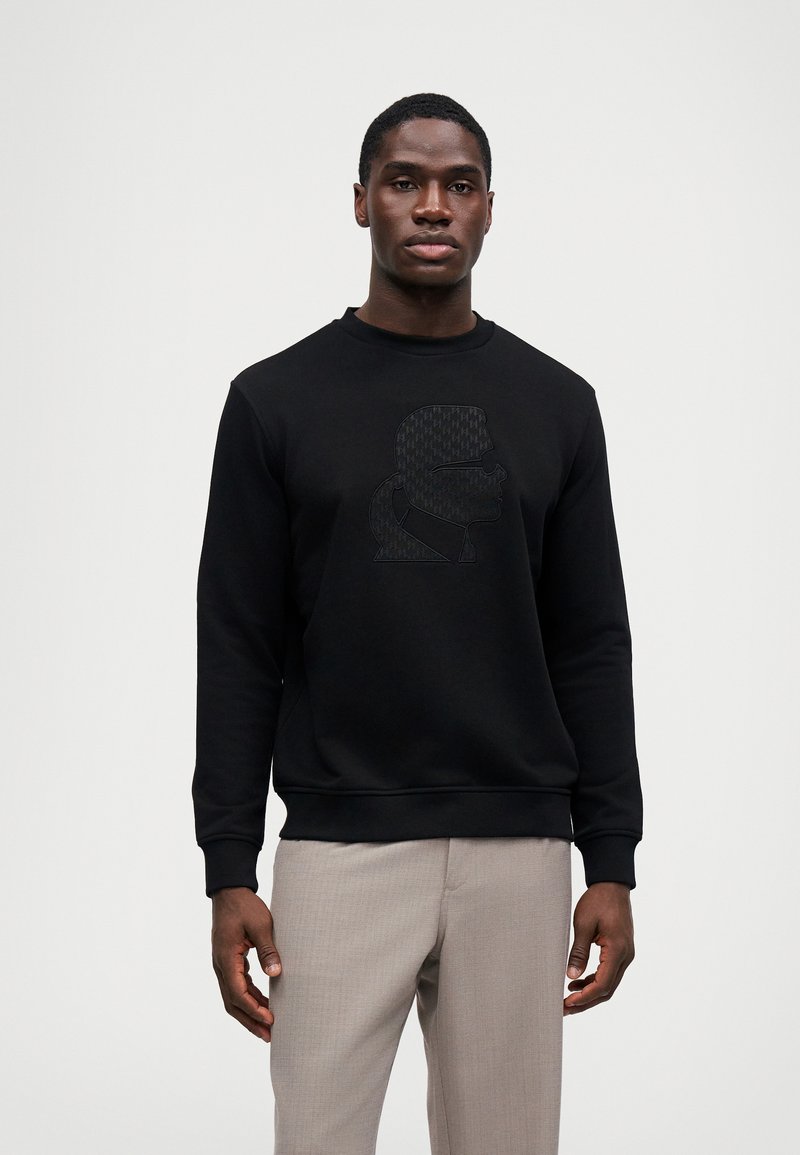 Μαύρη μπλούζα με κουκούλα που διαθέτει υφή στο γραφικό τύπωμα μπροστά, στρογγυλή λαιμόκοψη και ριμπ μανίκια. Το μοντέλο φοράει ανοιχτό γκρίζο παντελόνι.