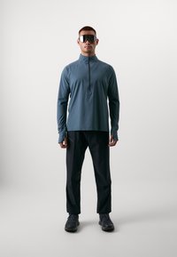 Prestatietop met lange mouwen in turquoise met een ritskraag, gecombineerd met zwarte broek en donkere schoenen. Vergezeld van grote reflecterende zonnebrillen.