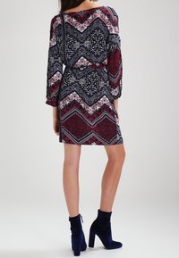 Kurzärmliges, gemustertes Kleid in Schwarz, Rot und Weiß mit geometrischen Designs. Kombiniert mit blauen, knöchelhohen, hochhackigen Stiefeln. Glatte Textur.