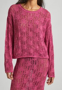 Różowy sweter z dzianiny o luźnym kroju, z koronkowym wzorem i długimi rękawami, zestawiony z pasującą spódnicą z tego samego materiału.