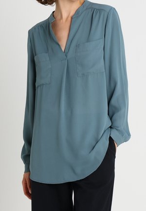 Woman wearing a long-sleeve teal blouse with two chest pockets, standing with one hand in black pants pocket against a plain background.