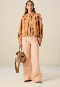 Veste courte marron clair avec des manches bouffantes, boutons noirs, portée avec un pantalon évasé rose pâle taille haute et un sac à main perforé beige.