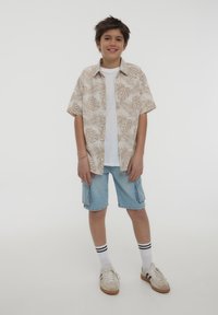 Camicetta beige a maniche corte con motivo a foglie di palma, t-shirt bianca sotto, pantaloni corti in denim e sneakers bianche con strisce nere.