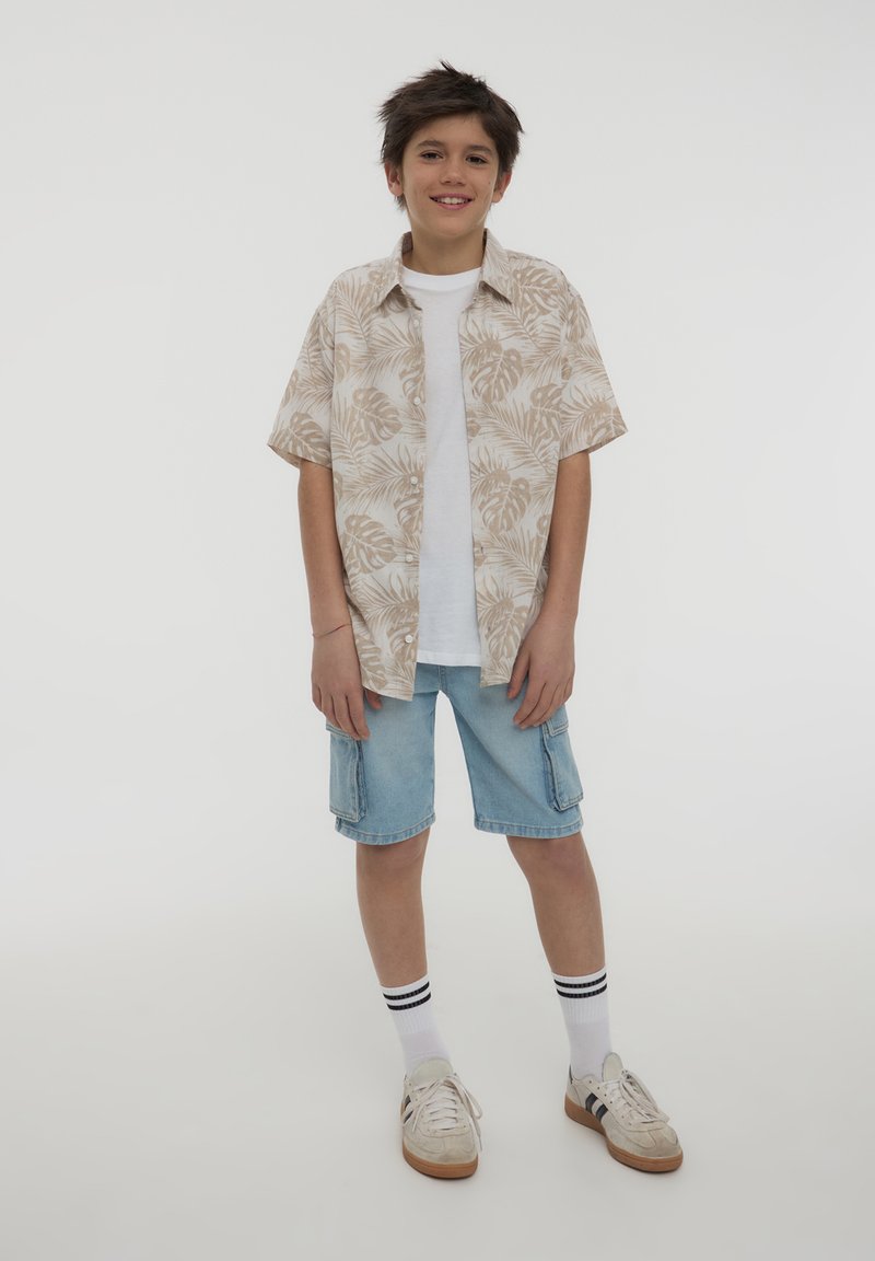 Camicetta beige a maniche corte con motivo a foglie di palma, t-shirt bianca sotto, pantaloni corti in denim e sneakers bianche con strisce nere.
