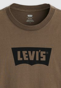 Ruda medvilninė marškinėliai su apvalia iškirpte. Ant jų yra didelis juodas „LEVI'S“ logotipas juodoje lenktoje formoje. Audinys turi tekstūrą.