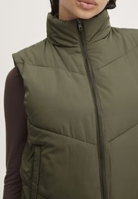 Gilet sans manches vert olive avec un col montant, fermeture éclair et motif de matelassage en chevrons. Dispose de poches latérales et d'une finition lisse et texturée.