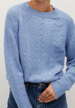 Jasnoniebieski sweter z dzianiny z wzorami chevron i warkoczy, noszony z ciemnoniebieskimi jeansami z wysokim stanem, ręka opiera się w pobliżu talii.