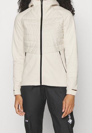 Osoba ubrana w jasnobeżową kurtkę z kapturem zapinaną na zamek, z pikowaną klatką piersiową i czarnymi kieszeniami na zamek, w połączeniu z czarnymi spodniami z białymi logo.