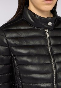 Giacca a vento nera con design trapuntato orizzontale, colletto alto e chiusura frontale con zip e dettagli in metallo argentato. Texture leggermente lucida.