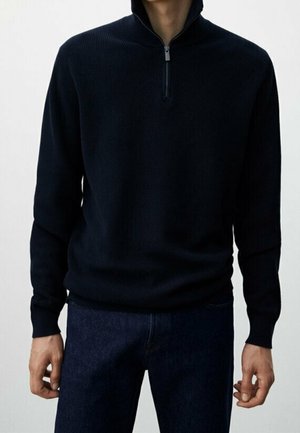 Homme portant un pull noir à demi-fermeture éclair et un jean bleu foncé, debout les bras détendus le long du corps devant un fond uni.