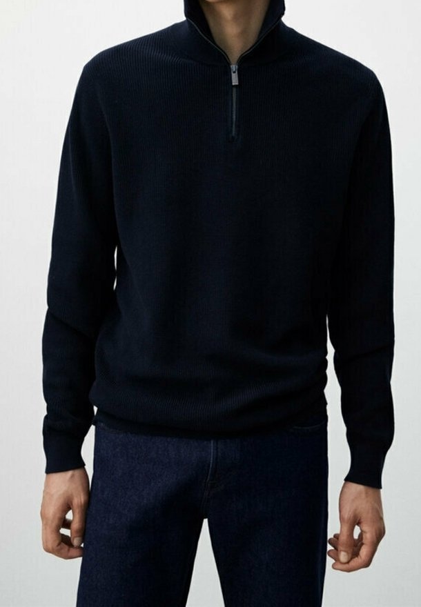 Homme portant un pull noir à demi-fermeture éclair et un jean bleu foncé, debout les bras détendus le long du corps devant un fond uni.