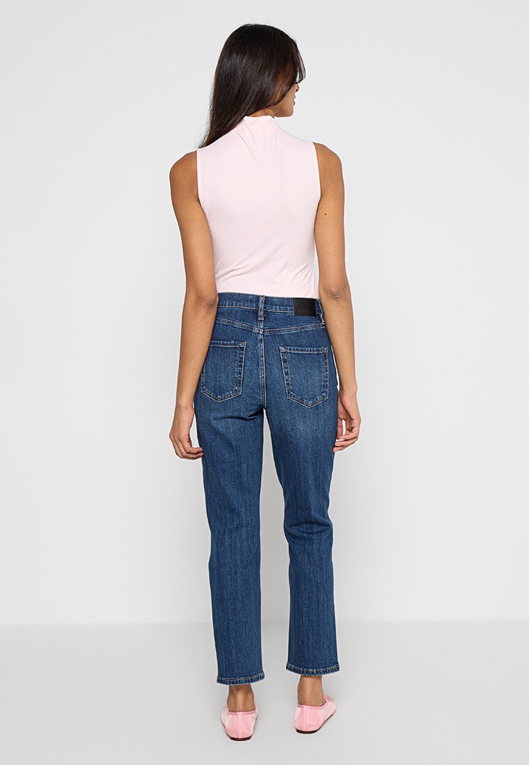 Rosa ärmelloses Top, blaue hochtaillierte Jeans mit zwei Gesäßtaschen und hellrosa flachen Schuhen. Glatter Stoff mit einer taillierten Silhouette.