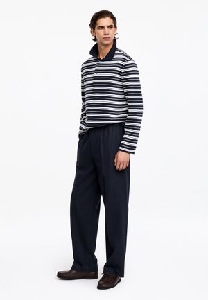 Jeune homme debout, portant un polo à manches longues rayé noir et blanc, un pantalon ample bleu marine foncé et des chaussures marron foncé.