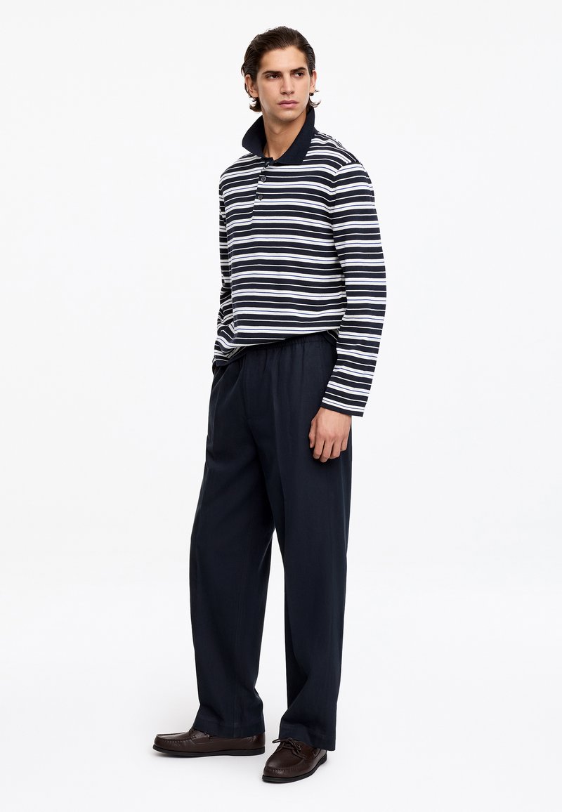 Jeune homme debout, portant un polo à manches longues rayé noir et blanc, un pantalon ample bleu marine foncé et des chaussures marron foncé.