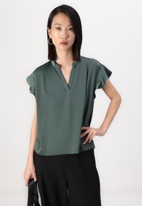 Dyp grønn bluse med korte volangermer, V-hals og glatt tekstur. Båret med svarte bukser. Minimalistisk design og ledig passform.