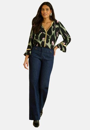 Woman wearing green, black, and pink patterned V-neck blouse with gold trim, dark blue high-waisted wide-leg jeans, and black shoes.