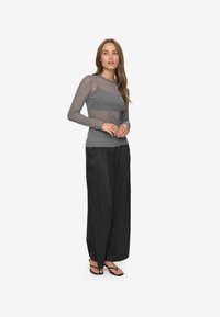 A hosszú ujjú, áttetsző, szürke topot viselő nő apró pöttyökkel, laza fekete széles szárú nadrágban és fekete, nyitott orrú szandálban áll, kezét összefonta.