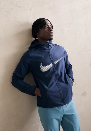 Marineblå vindjakke med hætte, prydet med et hvidt Nike-logo. Stoffet ser let og glat ud; modellen har blå bukser på.