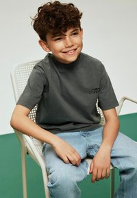 Gray short-sleeved T-shirt with a high neckline and subtle black logo, paired with light blue jeans. The model sits on a white chair.