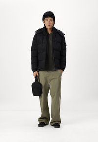 Schwarze Pufferjacke mit Kapuze, mit strukturierten Einsätzen, über einem dunklen Hemd getragen. Lockere olivgrüne Hose, hält eine kleine schwarze Tasche.