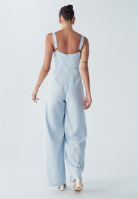 Svetlo modra kombinezona z debelimi naramnicami in nastavljivo kovinsko opremo, ki ima prilegajočo se zgornji del in široke, tekoče hlače, tekstura tkanine je gladka.