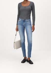 Graues Langarmoberteil mit Knopfdetails, hellblaue Skinny-Jeans und schwarze Slipper. Das Model trägt eine beige Handtasche.