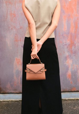 Small brown leather handbag with a geometric flap, gold hardware, and a top handle, held by a person wearing a gray top and black skirt.